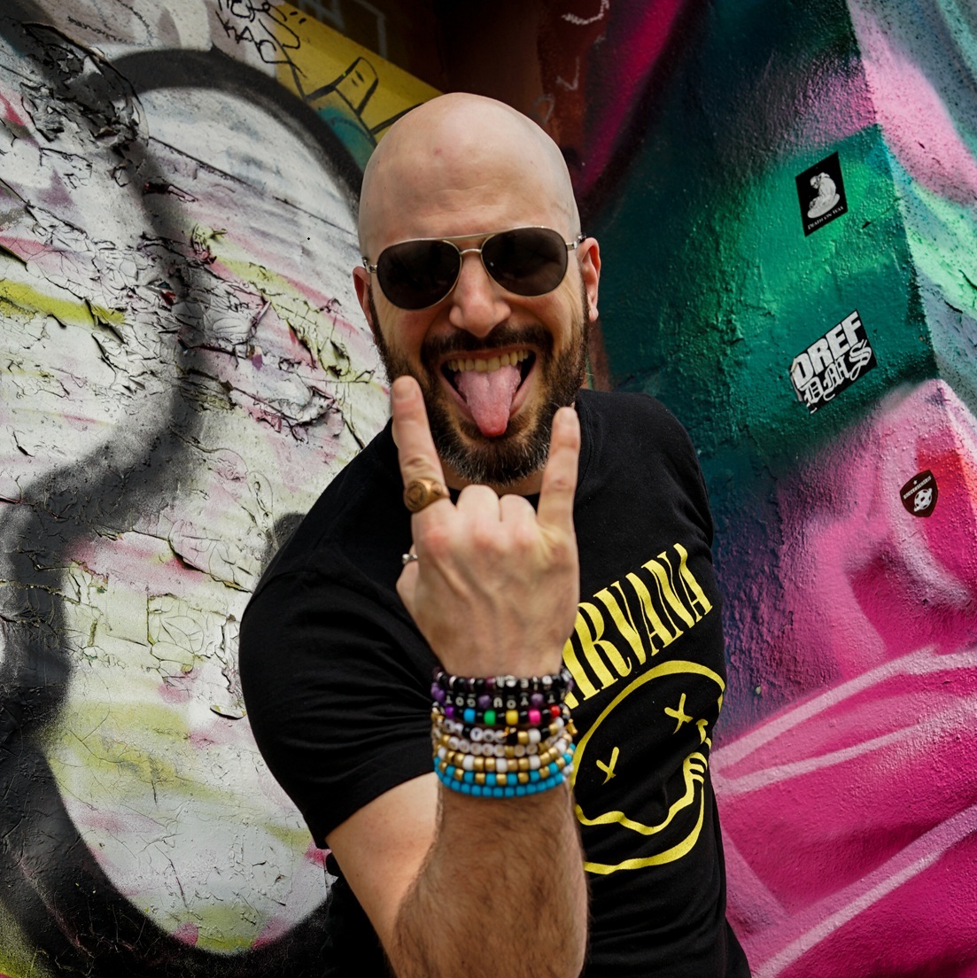Mark Pompeo AKA Mark Wise press shot in front of a building with graffiti. Wearing sunglasses, tongue sticking out, making the heavy metal horns symbol. Wearing a Nirvana t-shirt and rave kandi bracelets prominently shown on his arm.