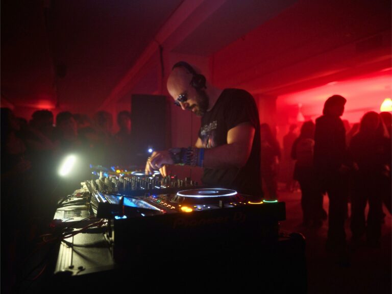 Company owner Mark Pompeo DJ'ing with a pair of CDJs and mixer in a room with red lights and members of the crowd in front and behind him.