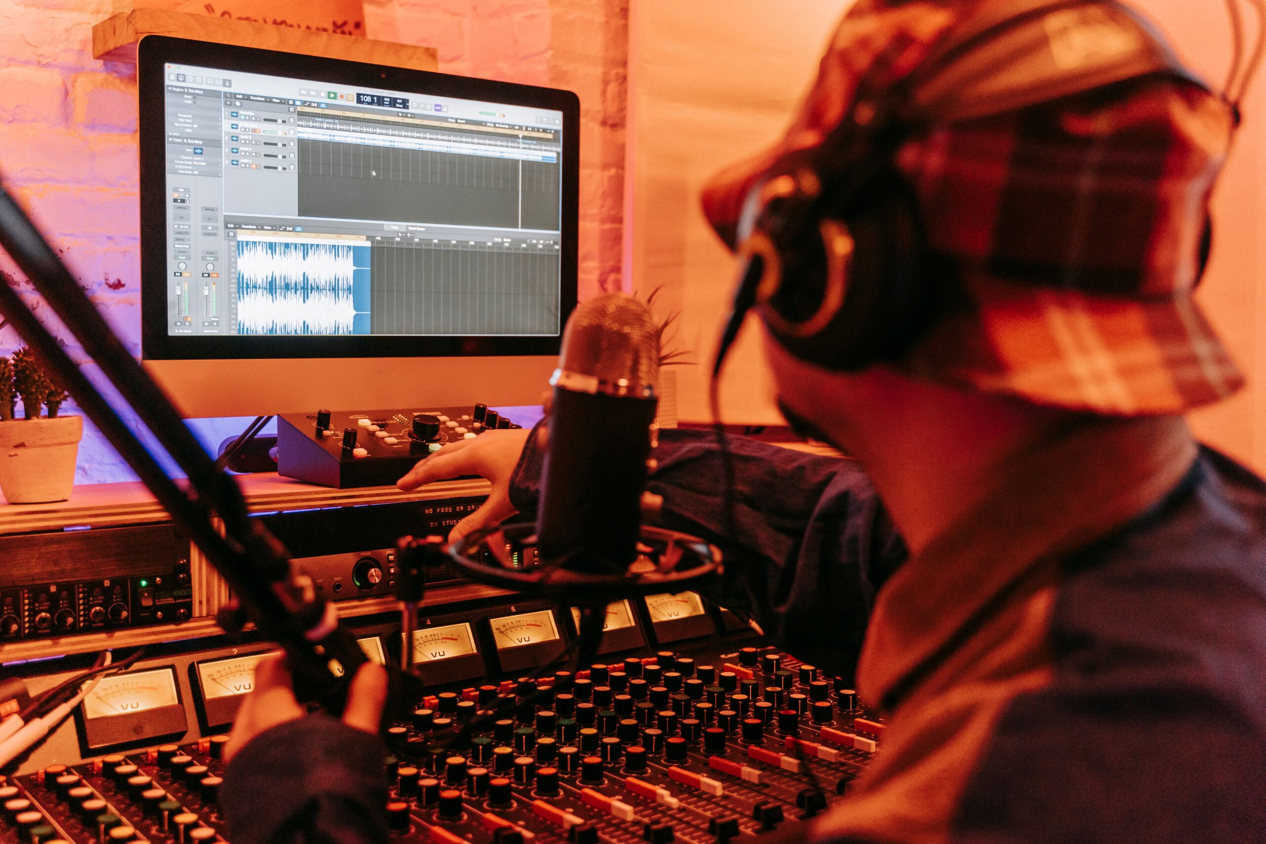 Music producer in a studio working on a song. Computer screen, microphone, and mixing board in front of them.