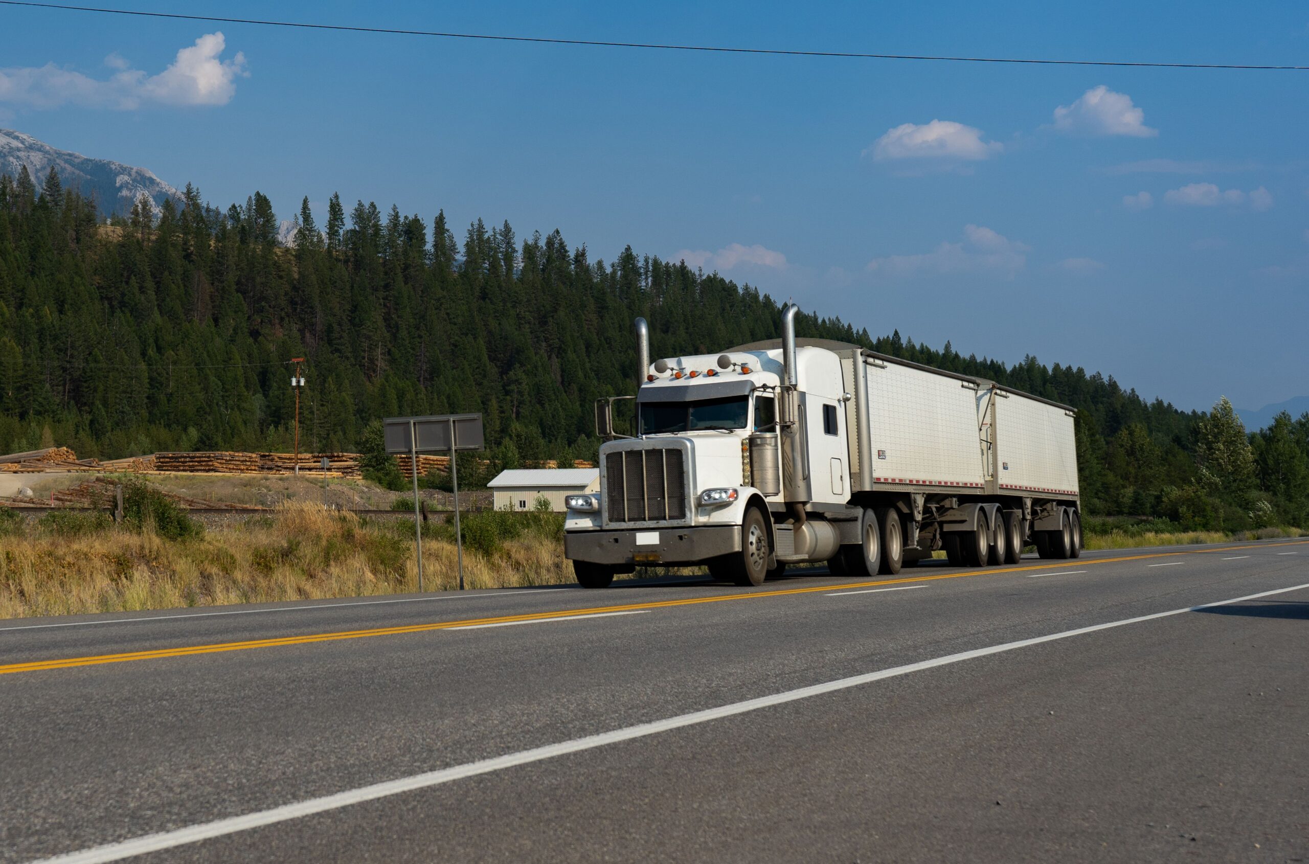 A white freight truck driving on tree-lined road with a blue sky and sparse clouds.
