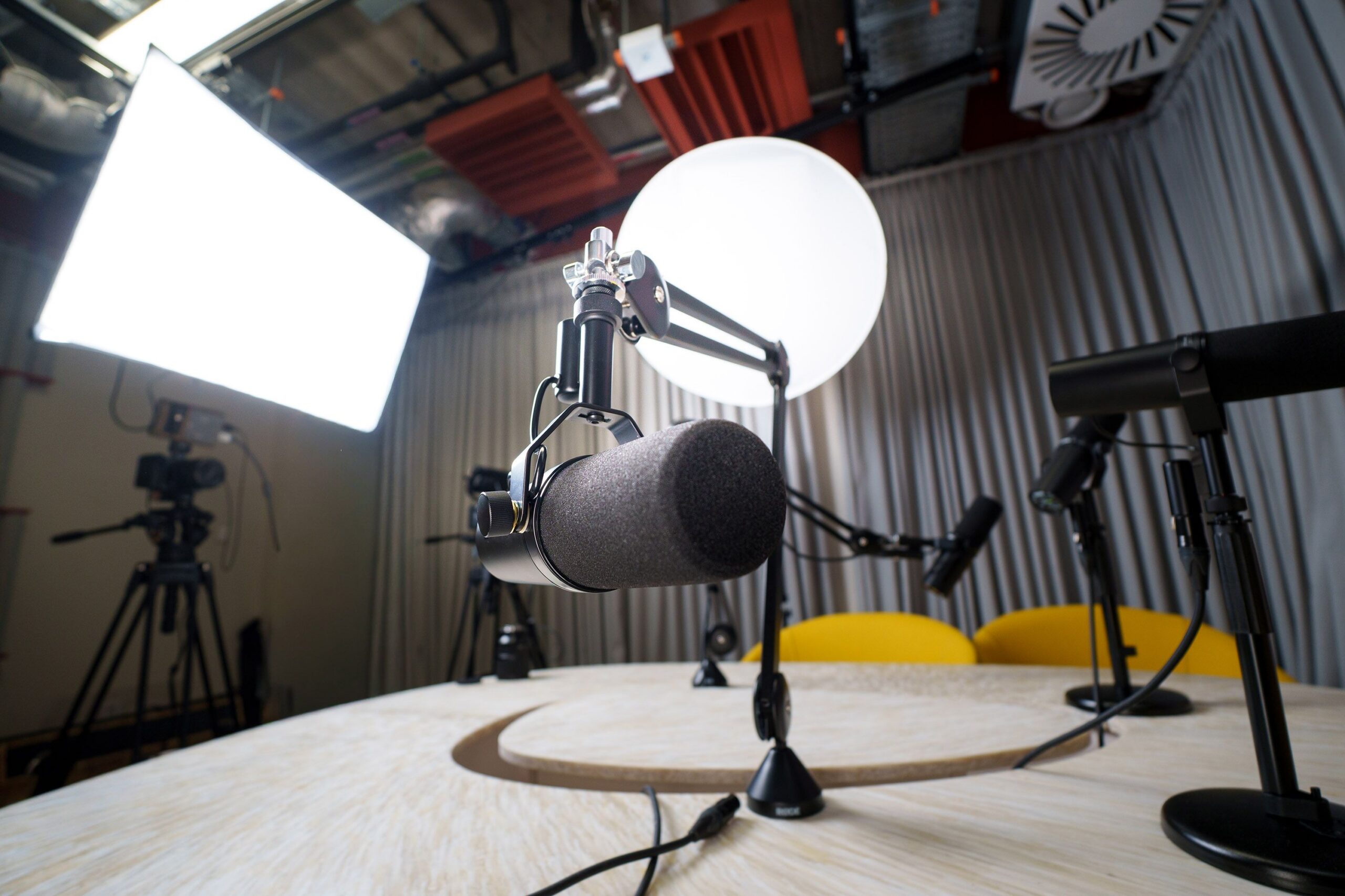 A social media content recording studio with two bright white lights, a cellphone and camera mounted beneath one of the lights, and microphones on a table with two yellow chairs, all surrounded by a gray curtain.