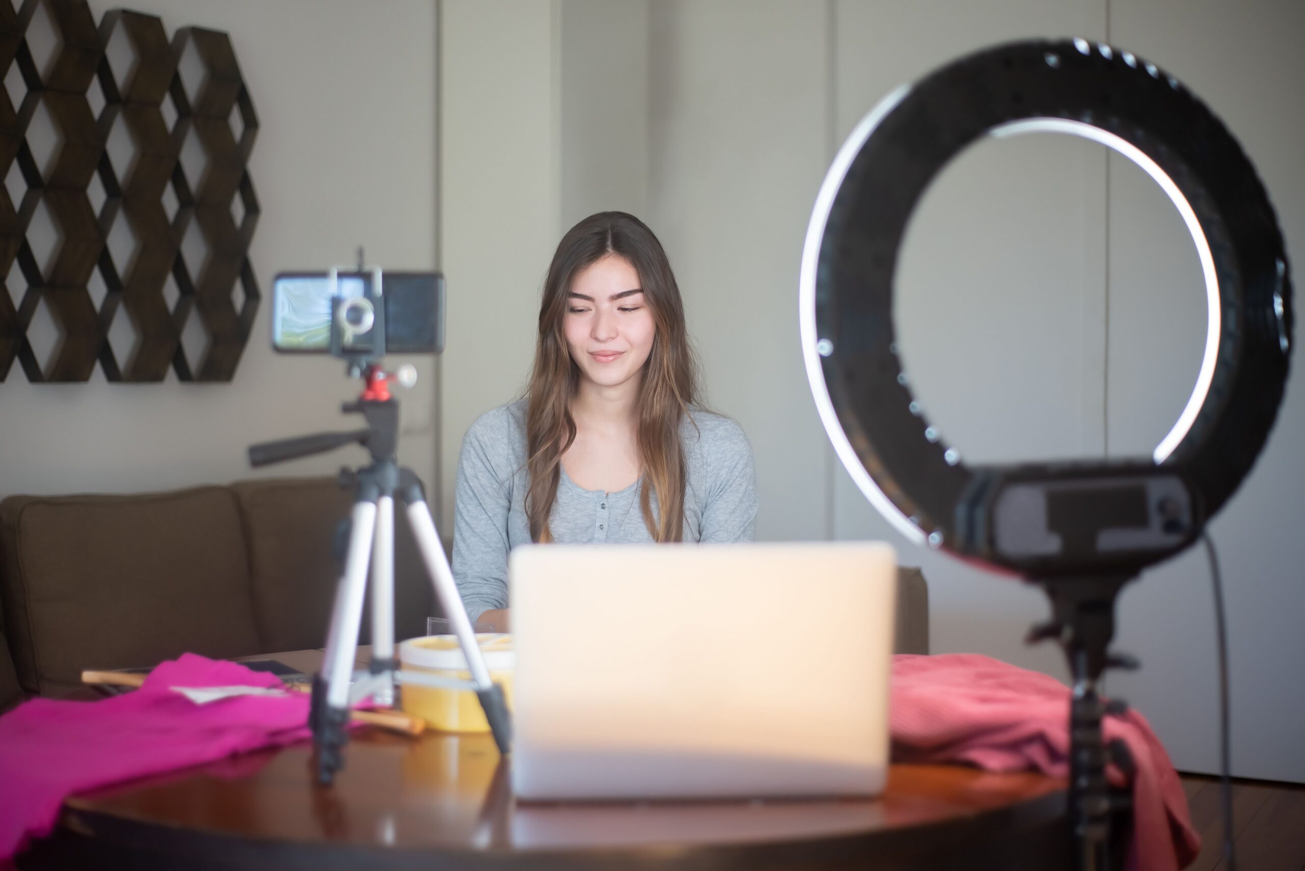 A woman sitting at a table with a laptop and clothing items filming video content with a cellphone and ring light.