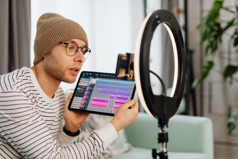 A man holding a tablet with audio software on the screen filming video content on a cellphone mounted to a ring light.