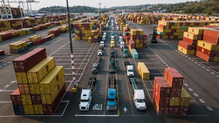 Three long lines of freight trucks at a port with stacks of shipping containers on both sides.