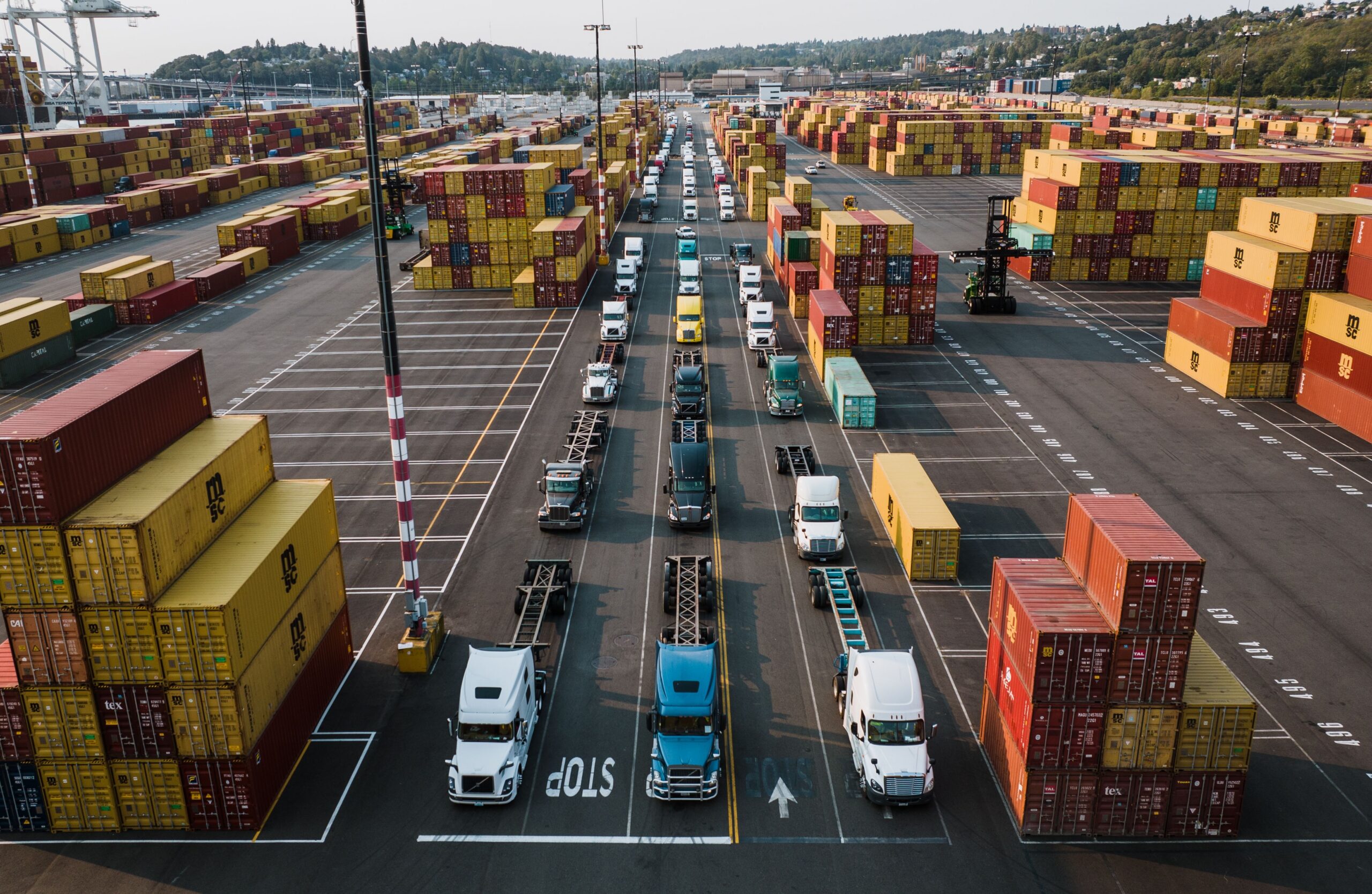 Three long lines of freight trucks at a port with stacks of shipping containers on both sides.