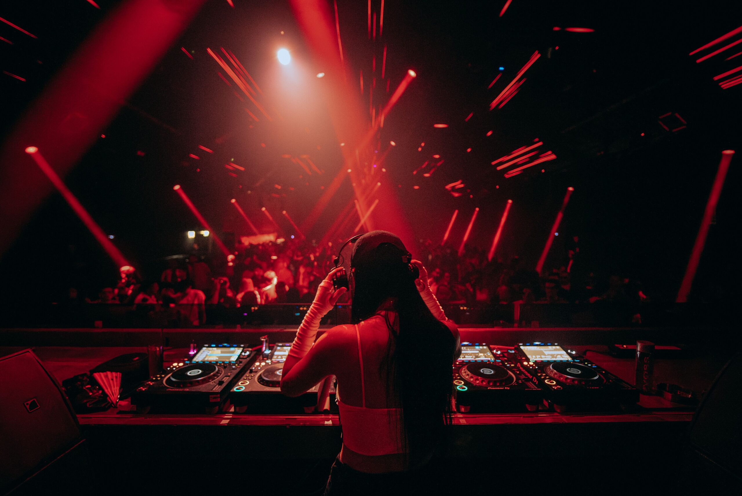 A DJ shot from behind in front of four CDJs performing at a rave in a club with many red lights projecting from the ceiling.