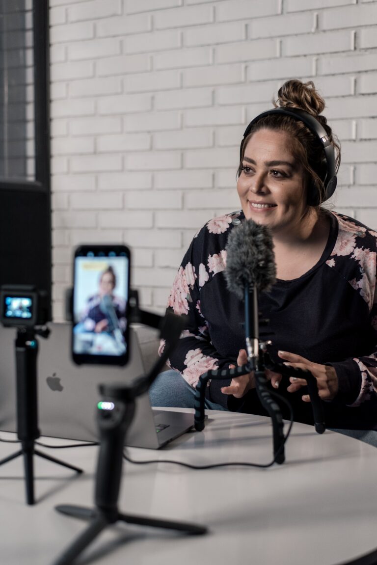 A woman recording social media content with a phone mounted in a stand, microphone in front of her face, and wearing headphones, all connected to a MacBook.