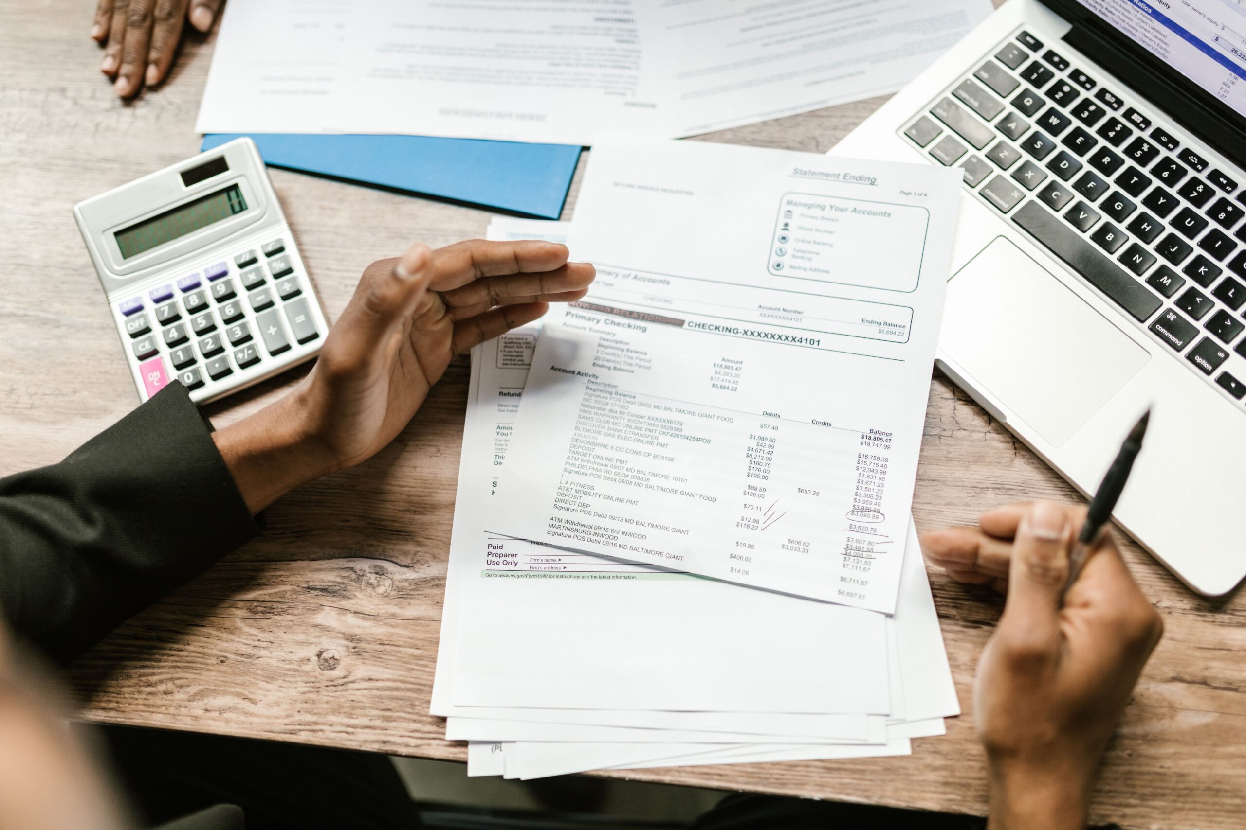 Person's hands at a desk with a laptop, calculator, and pages of business documents and reports. Right hand holding a pen.