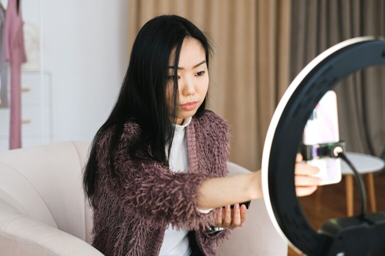 A woman sitting in a chair filming video content on a cellphone mounted to a ring light.