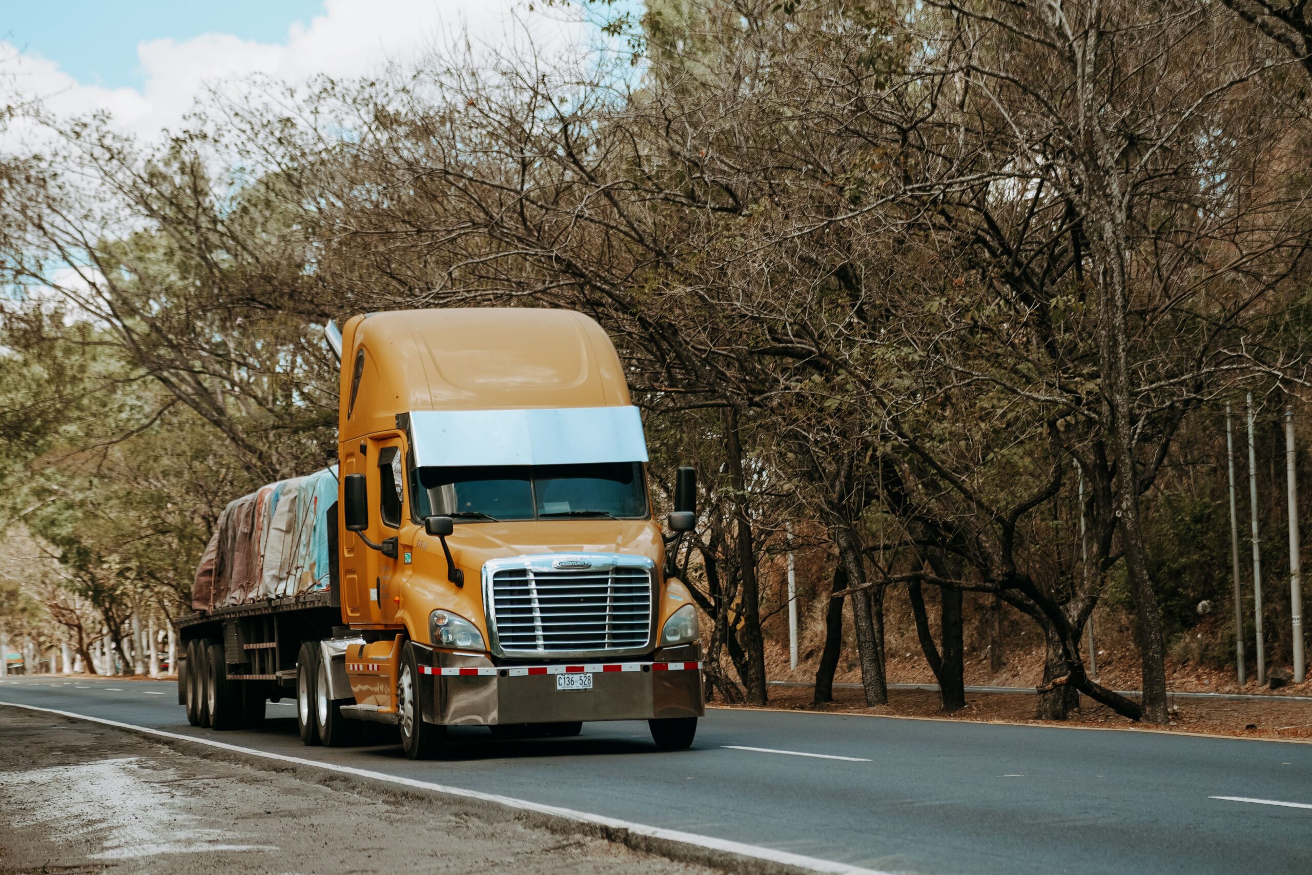 A flatbed freight truck with an orange cab driving on tree-lined road